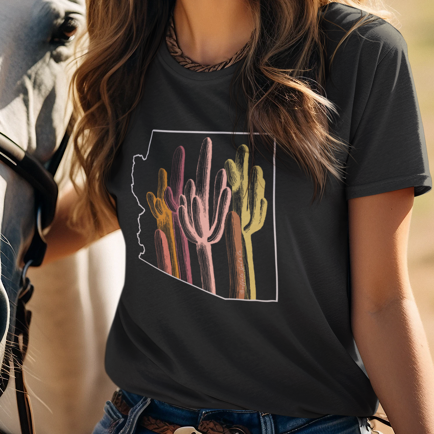 A smiling woman outdoors wearing a black graphic T-shirt with a colorful desert cactus design inside the state of Arizona outline, paired with a white cowboy hat and blue jeans, standing beside a horse.