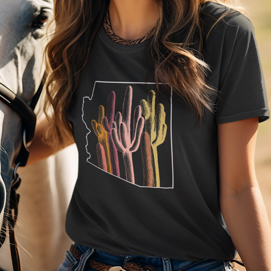 A smiling woman outdoors wearing a black graphic T-shirt with a colorful desert cactus design inside the state of Arizona outline, paired with a white cowboy hat and blue jeans, standing beside a horse.