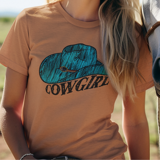 A smiling woman outdoors wearing a tan graphic T-shirt with a blue cowboy hat and the word 'COWGIRL' underneath, standing next to a white horse under a clear blue sky.