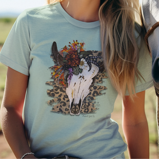 A smiling blonde woman stands outdoors next to a white horse, wearing a light blue graphic T-shirt with a horse skull and floral print. The background shows a bright blue sky and a grassy field.